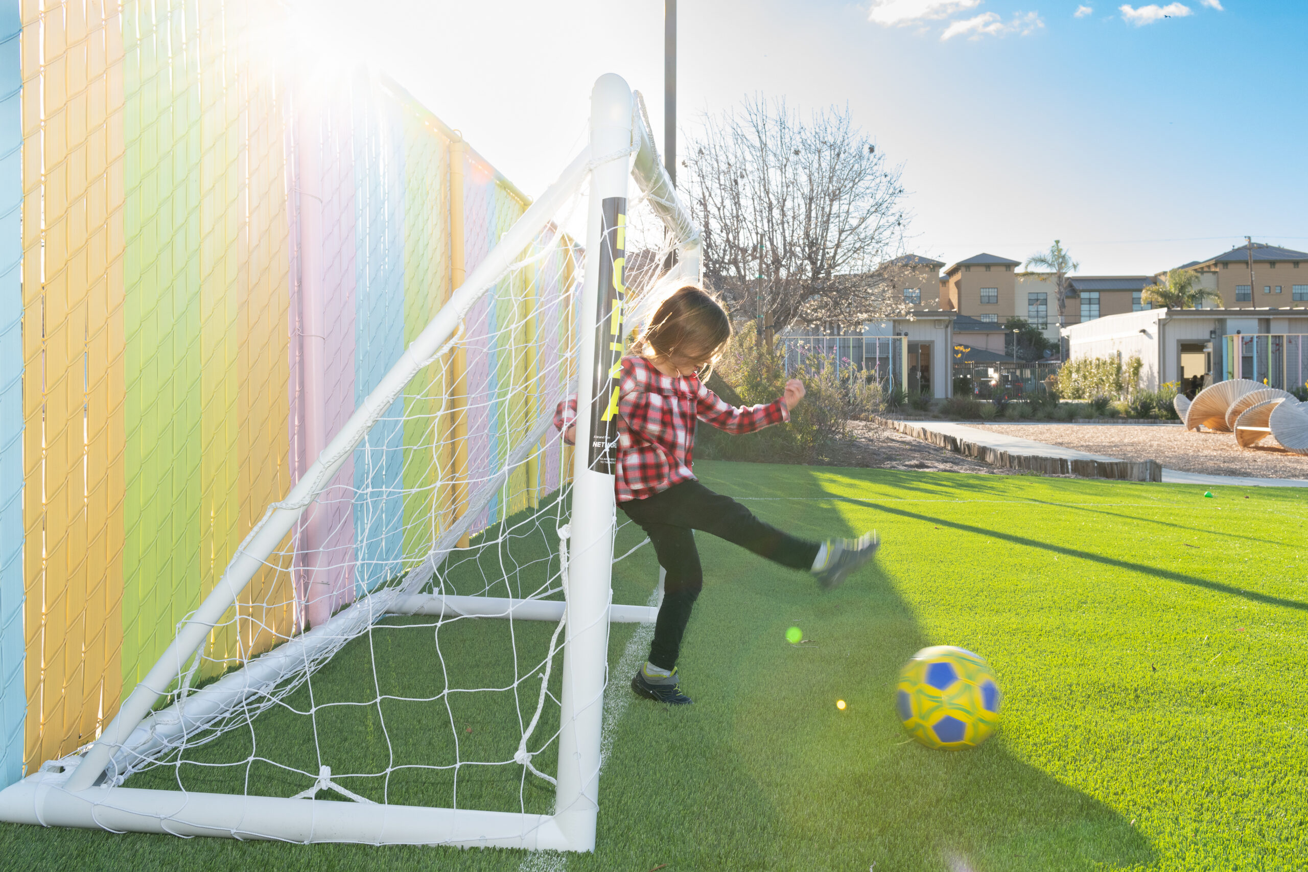 Child kicking a ball into a small goal on the turf yard beside a rainbow-painted fence