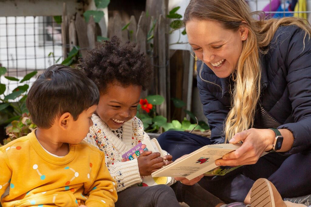 A teacher reading a picture book to two young children in a plant-filled corner