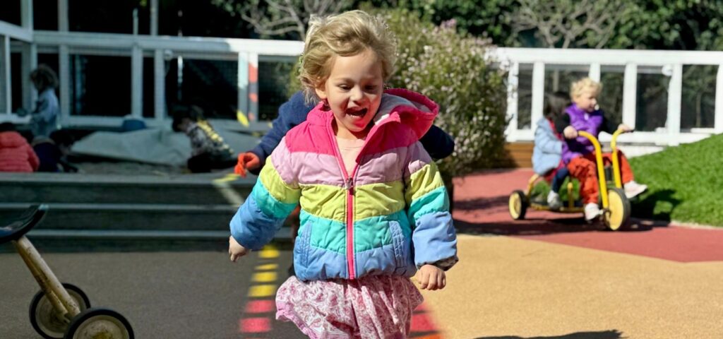 Preschooler in a rainbow jacket running across the painted play yard during camp
