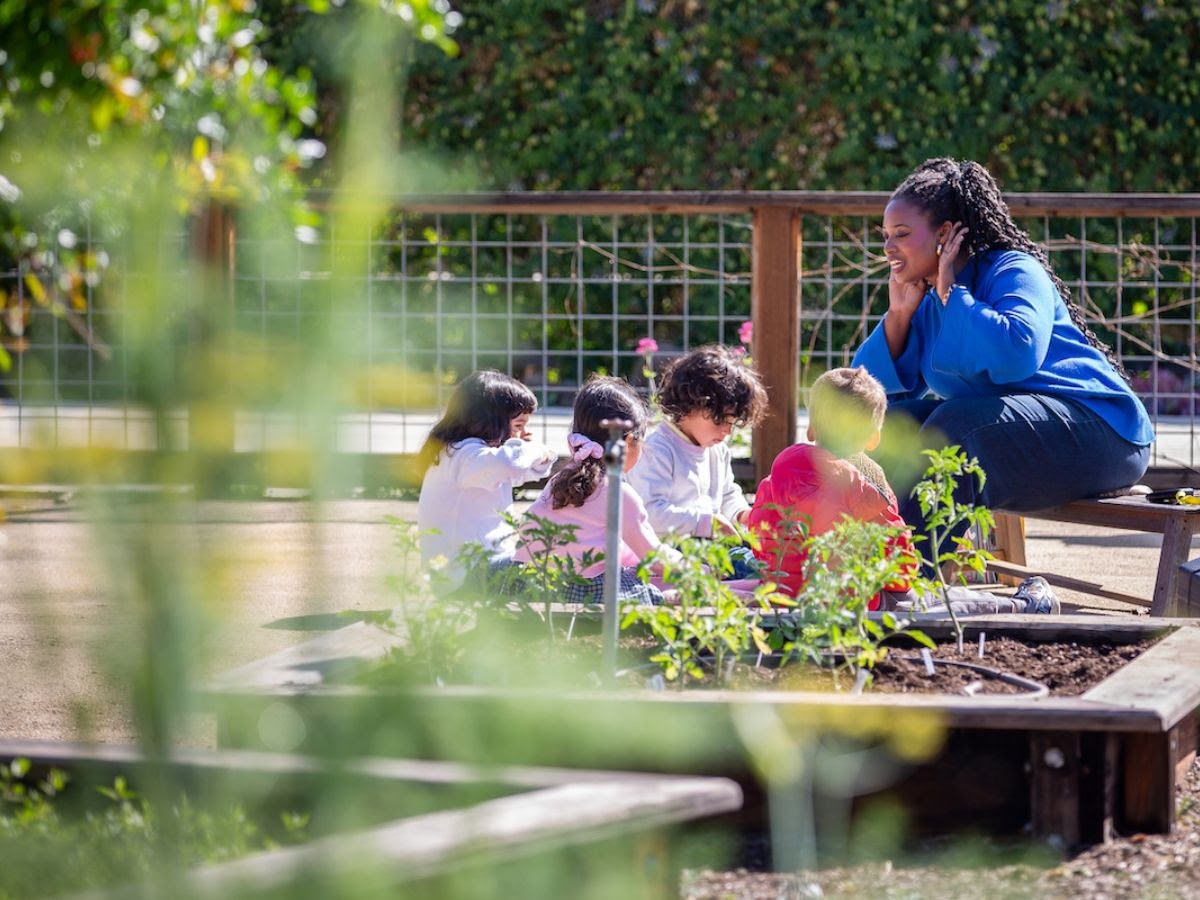 A La Scuola teacher crouches beside preschoolers planting in the raised garden beds.