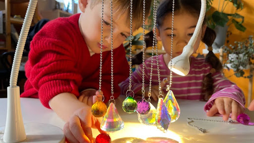 Children exploring hanging crystal prisms lit by a lamp on the atelier table