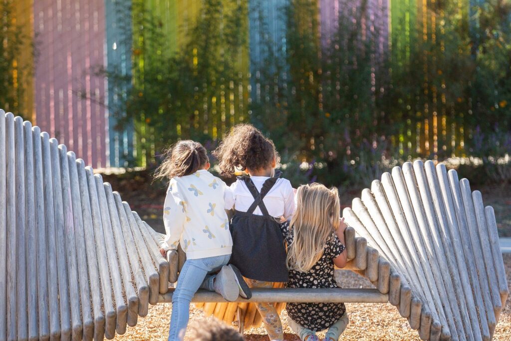 Three children sitting on a bench inside a curved wooden log structure