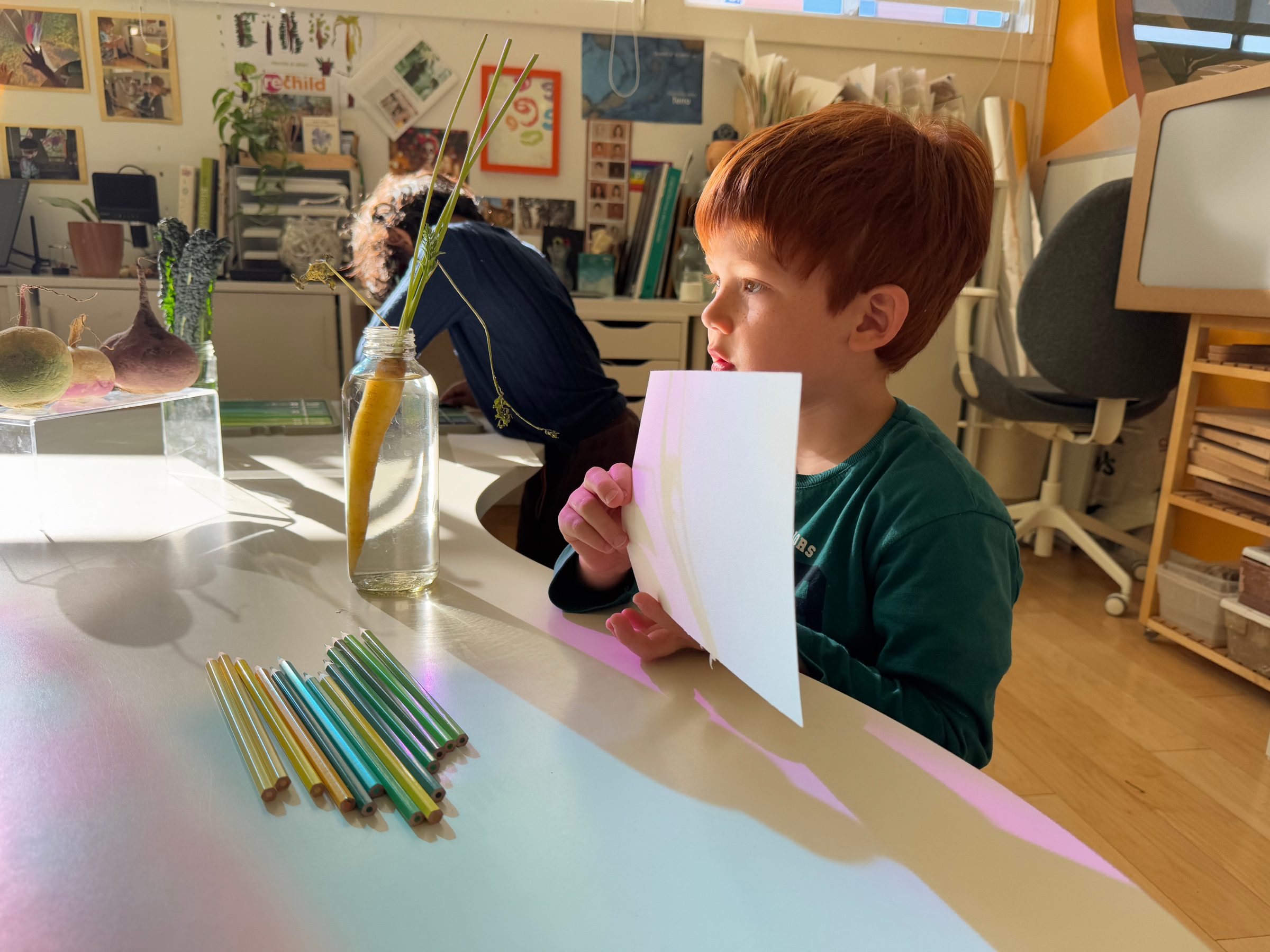 Child holding up a sheet of paper at a table strewn with colored pencils