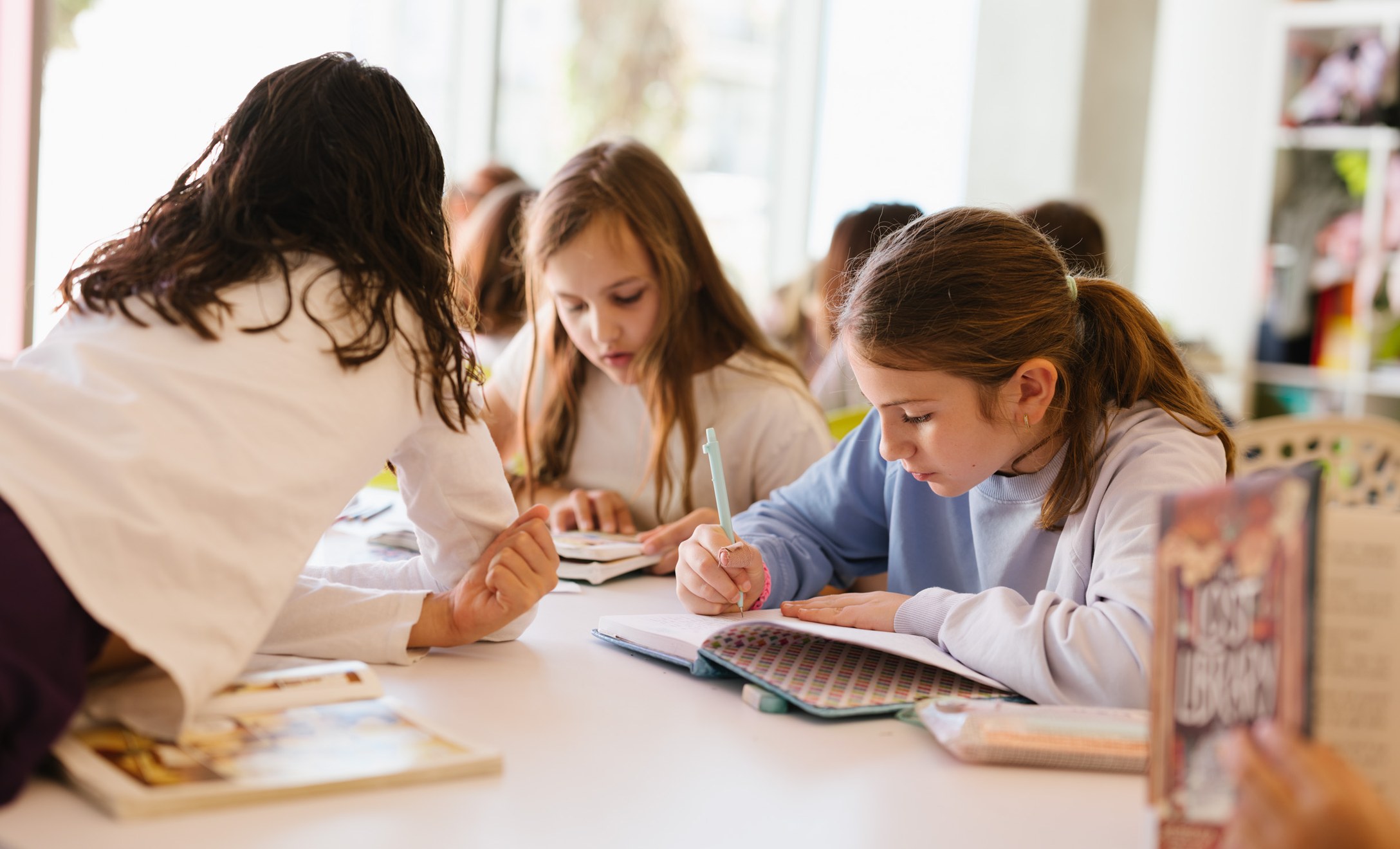Three elementary students writing in workbooks at a table while a teacher leans in to help