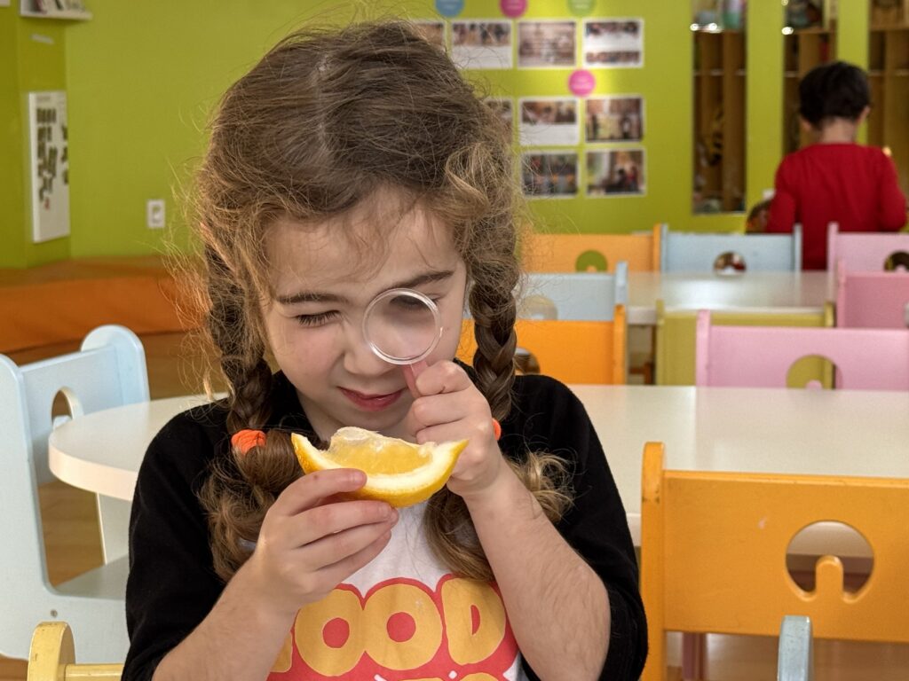 A girl looking through a magnifying glass while tasting an orange slice