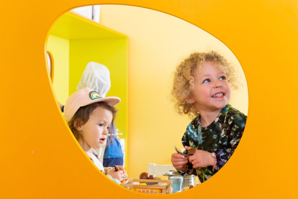 Two preschoolers peeking out of a round window in a bright yellow play house