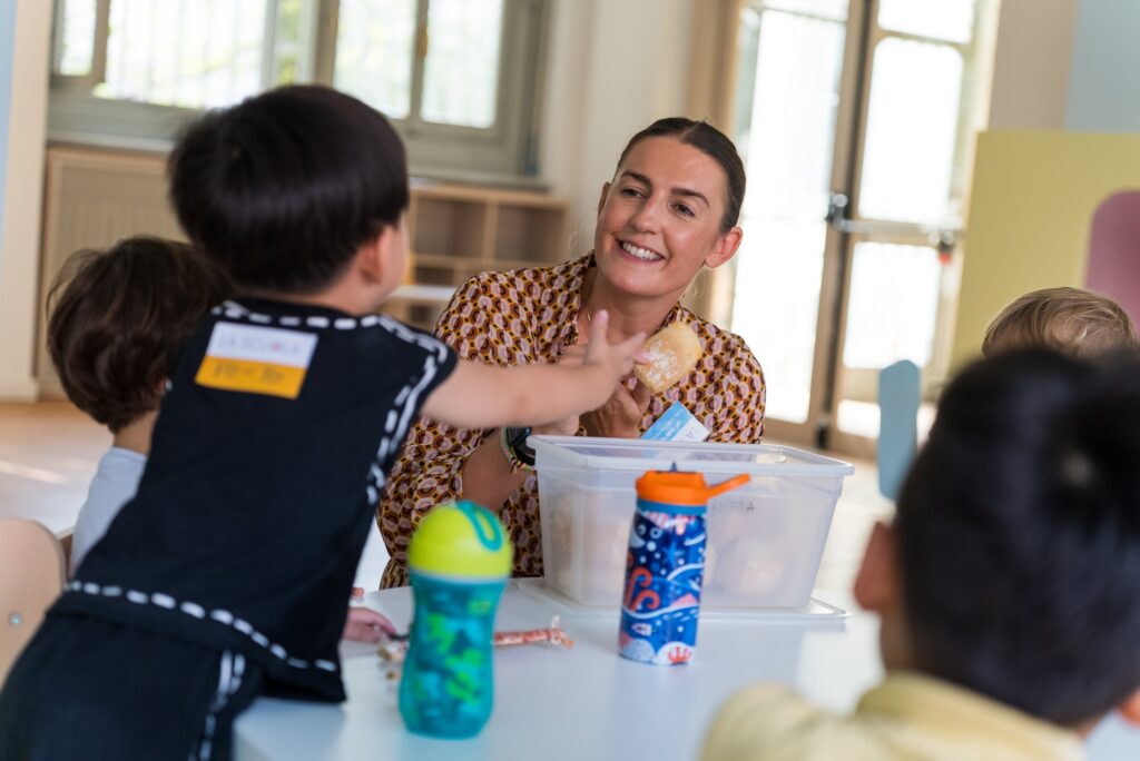 A preschool teacher smiling with children around a classroom table