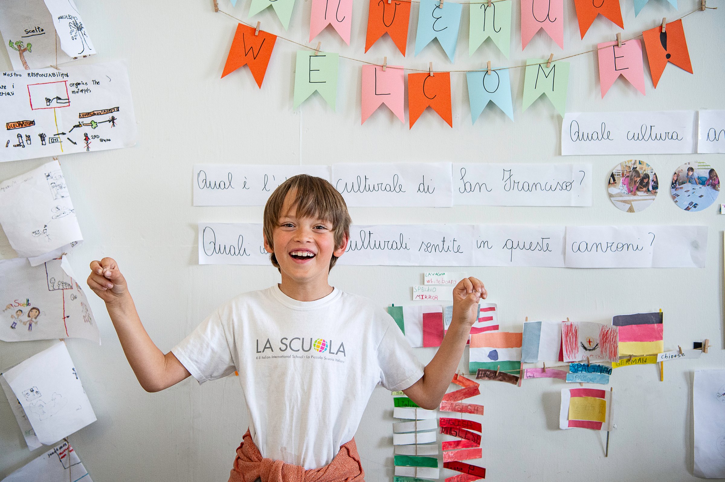 A smiling student in a La Scuola T-shirt in front of a wall of flags and Italian writing