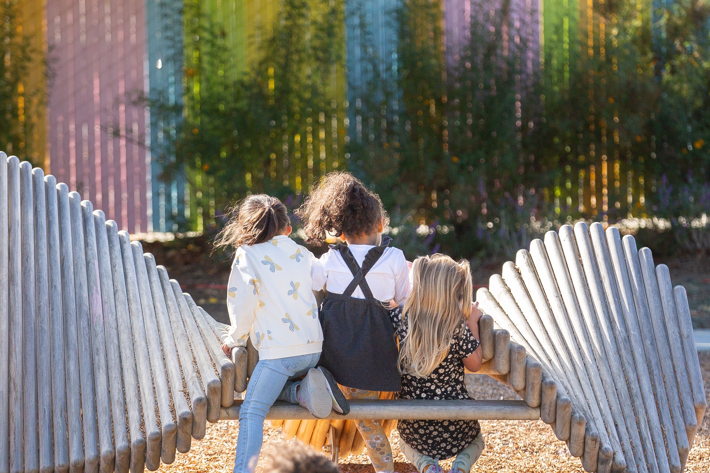 Three children sitting together on a carved wooden log bench in front of a pastel-painted fence